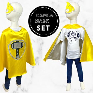 K&ouml;nnte beinhalten: Gelbes Superhelden-Umhang- und Maskenset f&uuml;r Kinder. Der Umhang hat einen silbernen Schild mit einem schwarzen Hammer. Die Maske hat ein gelbes Design. Das T-Shirt sagt "Sei dein eigener Held".