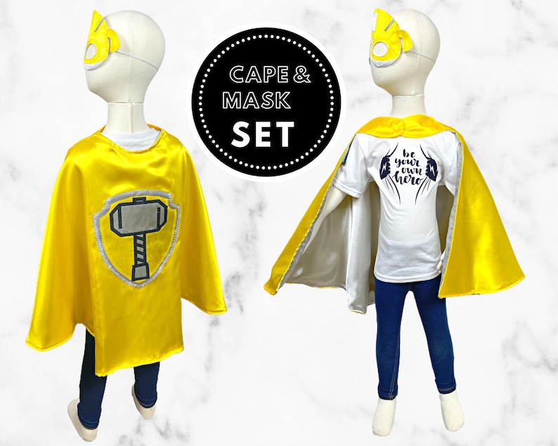 K&ouml;nnte beinhalten: Gelbes Superhelden-Umhang- und Maskenset f&uuml;r Kinder. Der Umhang hat einen silbernen Schild mit einem schwarzen Hammer. Die Maske hat ein gelbes Design. Das T-Shirt sagt "Sei dein eigener Held".