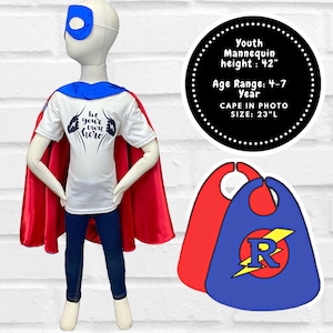 May include: A mannequin dressed in a superhero costume, including a white t-shirt with the text "be your own hero", a red cape, blue jeans, and a blue eye mask. The cape is 23 inches long. The mannequin is 42 inches tall and is for ages 4-7.
