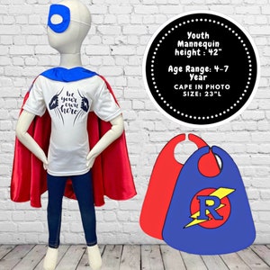 May include: A white t-shirt with the text "Be your own hero" printed on it. A red cape is draped over the shoulders of a mannequin wearing the shirt. The mannequin is wearing blue jeans and a blue eye mask. Two capes are shown on the right side of the image. One cape is red and the other is blue with a yellow lightning bolt and the letter "R" in a red circle. The text "Youth Mannequin height: 42" Age Range: 4-7 Year CAPE IN PHOTO SIZE: 23"L" is printed in white on a black circle.