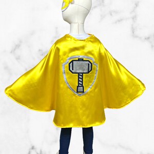 K&ouml;nnte beinhalten: Gelber Superhelden-Umhang mit einem silbernen Schild und einem Hammer-Design. Der Umhang ist aus Satin und hat eine spitze Kapuze.