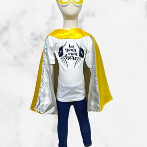 K&ouml;nnte beinhalten: Ein Superhelden-Kost&uuml;m f&uuml;r Kinder mit einem gelben Umhang, einer gelben Maske mit wei&szlig;em Visier und einem wei&szlig;en T-Shirt mit dem Text "be your own hero" in Schwarz.