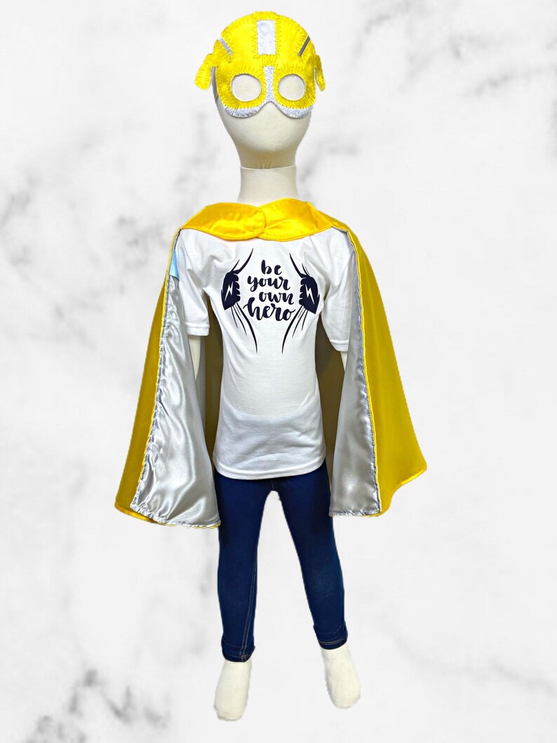 K&ouml;nnte beinhalten: Ein Superhelden-Kost&uuml;m f&uuml;r Kinder mit einem gelben Umhang, einer gelben Maske mit wei&szlig;em Visier und einem wei&szlig;en T-Shirt mit dem Text "be your own hero" in Schwarz.
