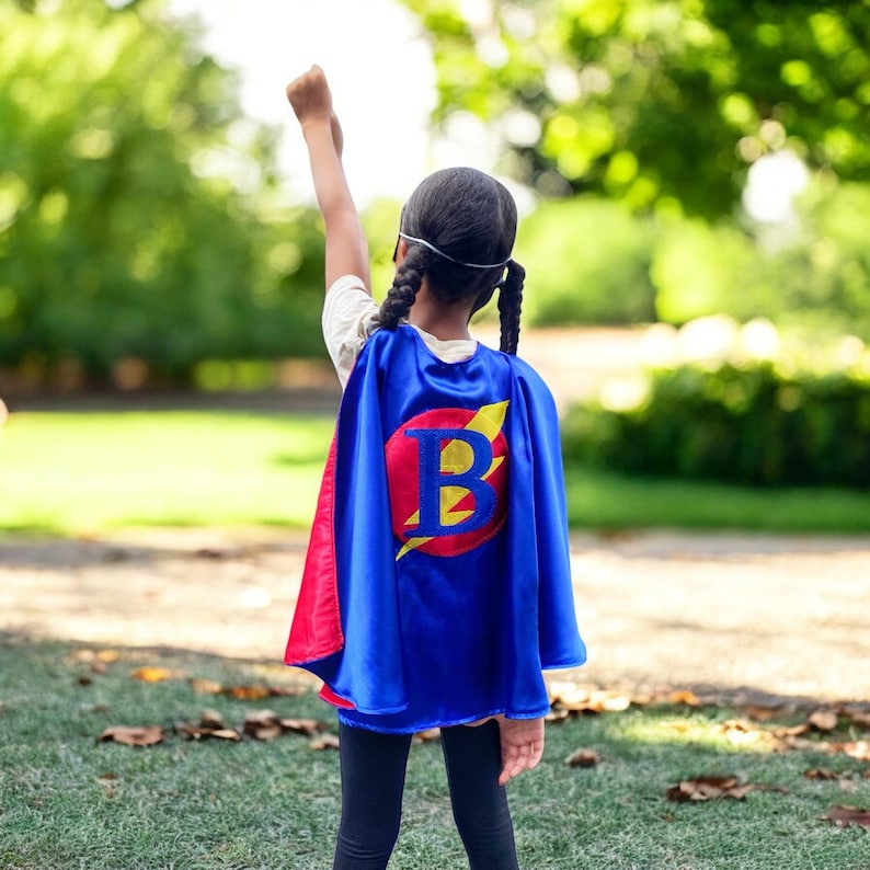 Peut inclure: Un enfant portant une cape de super-h&eacute;ros bleue et rouge avec un &eacute;clair jaune et la lettre "B" brod&eacute;e sur le devant. La cape est port&eacute;e sur une chemise blanche et un pantalon noir.