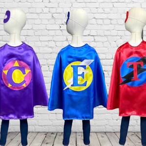 Peut inclure: Trois capes de super-h&eacute;ros de couleurs et de lettres diff&eacute;rentes. Les capes sont violettes, bleues et rouges. Les lettres sur les capes sont C, E et T. Chaque cape a un cercle jaune avec un &eacute;clair et une lettre au centre.