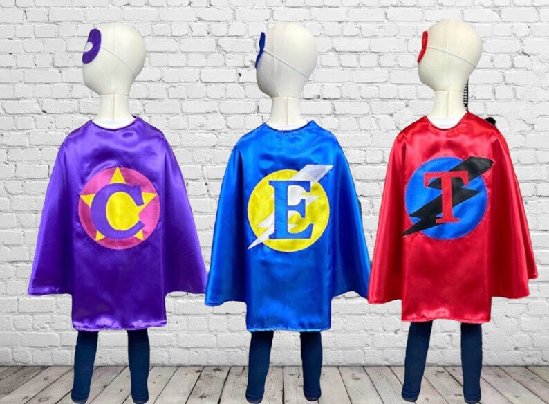 Peut inclure: Trois capes de super-h&eacute;ros de couleurs et de lettres diff&eacute;rentes. Les capes sont violettes, bleues et rouges. Les lettres sur les capes sont C, E et T. Chaque cape a un cercle jaune avec un &eacute;clair et une lettre au centre.