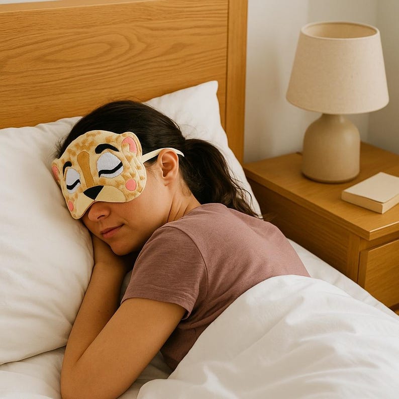 May include: A plush sleep mask shaped like a leopard face with embroidered closed eyes, a black nose, and rounded ears. The mask features a tan and brown spotted pattern with pink accents on the cheeks and ears, held in place by a white elastic strap.