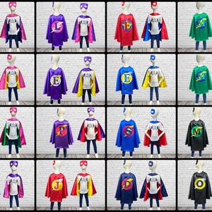 May include: A collection of superhero costumes for children, featuring capes, masks, and shirts. The capes come in various colors, including pink, red, blue, green, and black, each with a letter or symbol. The shirts are white with a printed design.