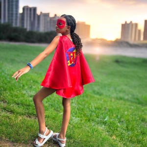 May include: A child wearing a red superhero cape and mask, with a circular emblem on the back. The emblem features a blue circle with a yellow and red design. The child is wearing white sandals and a blue watch, standing on green grass.