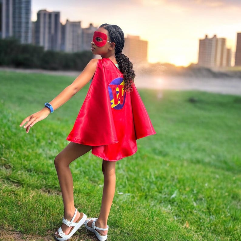 May include: A child wearing a red superhero cape and mask, with a circular emblem on the back. The emblem features a blue circle with a yellow and red design. The child is wearing white sandals and a blue watch, standing on green grass.