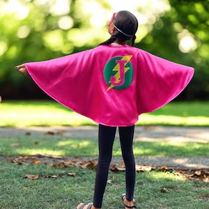 May include: A pink superhero cape with a green circle and a yellow lightning bolt. The circle has a pink letter "J" in the centre.