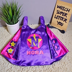 May include: A purple and pink superhero cape with the name "Nora" and the letter "N" in a starburst design. The cape has arm straps and two star patches. A wooden sign reads "Which Super Letter You".