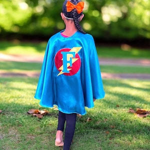May include: A child wearing a blue superhero cape with a red circle and a yellow lightning bolt. The circle has a large white letter "F" in the center. The child has a braid in their hair with an orange bow.