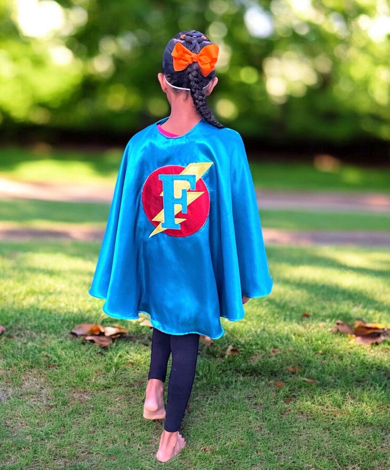 May include: A child wearing a blue superhero cape with a red circle and a yellow lightning bolt. The circle has a large white letter "F" in the center. The child has a braid in their hair with an orange bow.