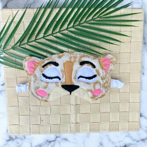 May include: A sleep mask shaped like a leopard face with a tan and brown spotted pattern, white closed eyes with black eyelashes, pink inner ears, pink cheeks, a tan nose bridge, and a black nose. The mask features a white elastic strap.
