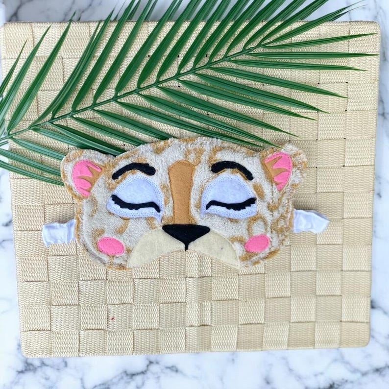 May include: A sleep mask shaped like a leopard face with a tan and brown spotted pattern, white closed eyes with black eyelashes, pink inner ears, pink cheeks, a tan nose bridge, and a black nose. The mask features a white elastic strap.