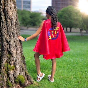May include: A red superhero cape with a circular emblem featuring a purple circle, a yellow lightning bolt, and the letter "W". The cape is worn by a person standing on green grass, with a tree and buildings in the background.