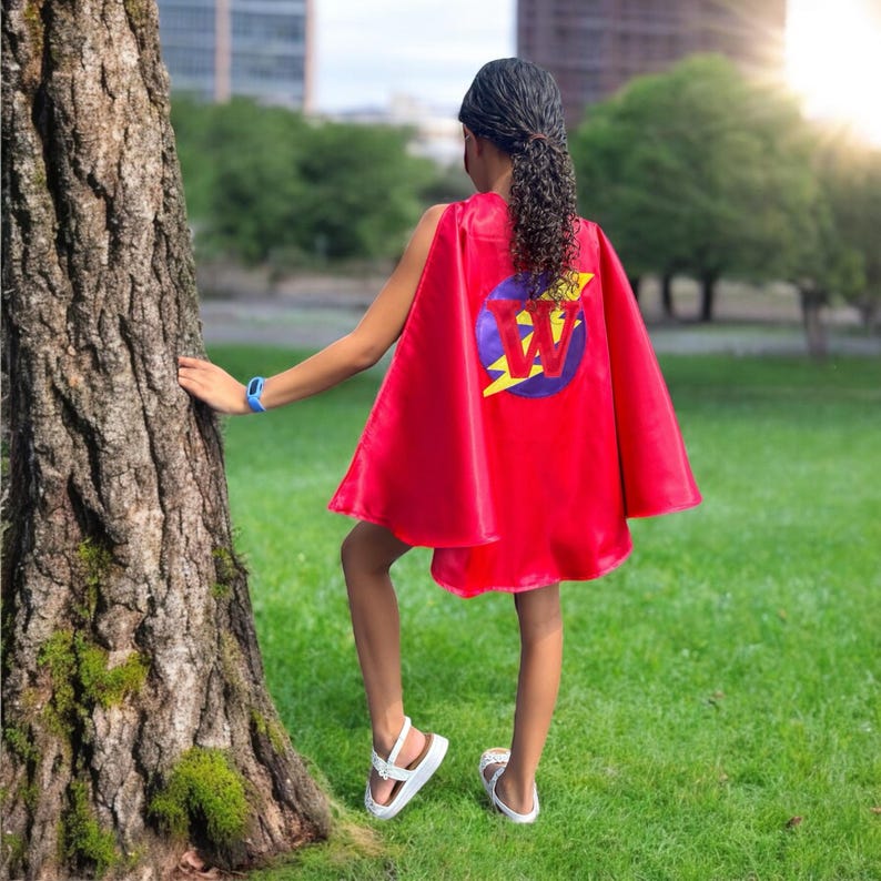 May include: A red superhero cape with a circular emblem featuring a purple circle, a yellow lightning bolt, and the letter "W". The cape is worn by a person standing on green grass, with a tree and buildings in the background.