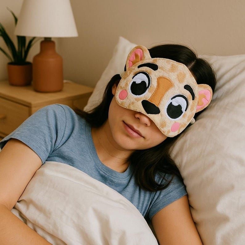 May include: A plush sleep mask shaped like a cartoon leopard face with tan fur, large black and white eyes, a brown nose, and pink cheek accents. The mask features rounded ears with pink inner details and covers the eyes with a soft, fuzzy texture.