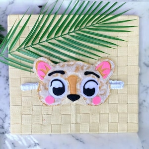 May include: A plush sleep mask shaped like a stylized leopard face with a tan and brown spotted pattern, large white eyes with black pupils, a tan nose, and bright pink accents on the ears and cheeks, featuring a white elastic head strap.