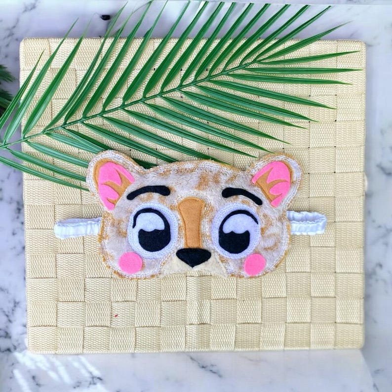 May include: A plush sleep mask shaped like a stylized leopard face with a tan and brown spotted pattern, large white eyes with black pupils, a tan nose, and bright pink accents on the ears and cheeks, featuring a white elastic head strap.