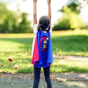 Peut inclure: Une jeune personne portant une cape de super-h&eacute;ros bleue et rose avec une lettre jaune "B" au dos, debout les bras lev&eacute;s en l'air, &agrave; l'ext&eacute;rieur dans un espace herbeux.