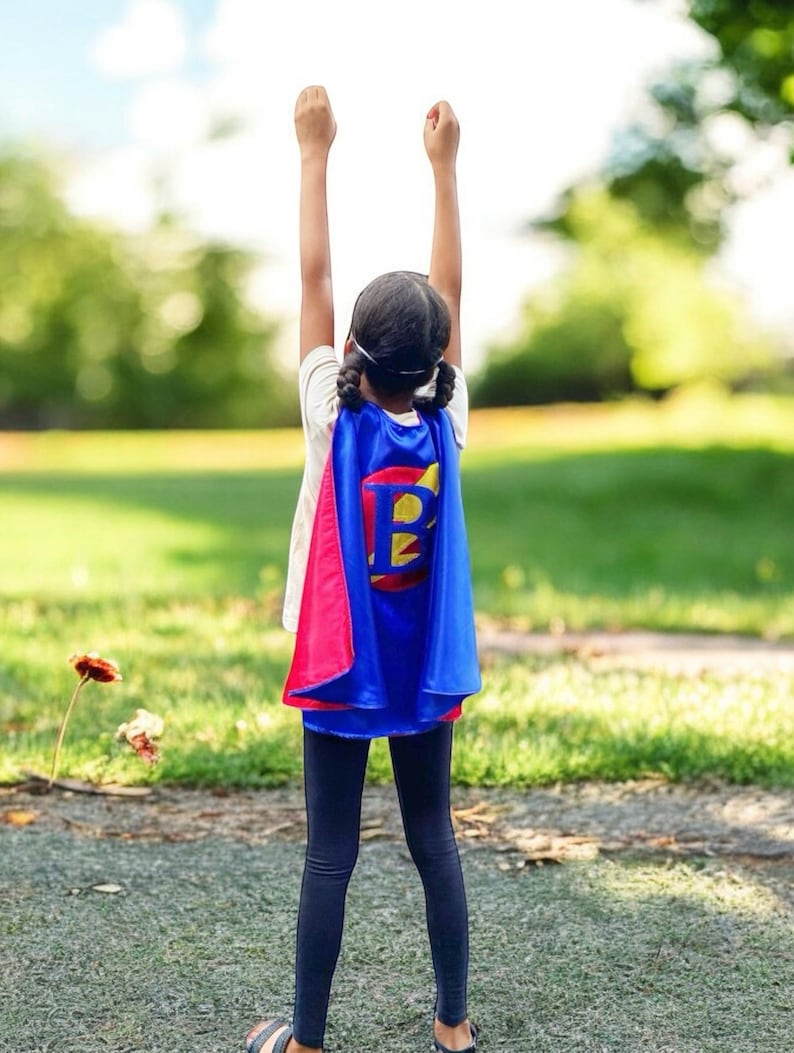 Peut inclure: Une jeune personne portant une cape de super-h&eacute;ros bleue et rose avec une lettre jaune "B" au dos, debout les bras lev&eacute;s en l'air, &agrave; l'ext&eacute;rieur dans un espace herbeux.