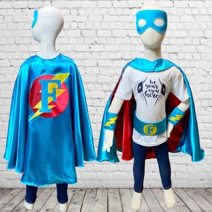 May include: A child's superhero costume set with a blue cape, red and yellow lightning bolt design, a white t-shirt with the text "Be your own hero", a blue mask, and a yellow belt with the letter "F" on it.