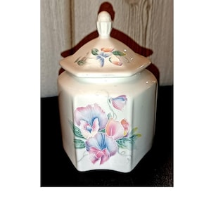 May include: A white ceramic jar with a lid, featuring a floral design in shades of pink, blue, and purple. The jar has an octagonal shape and a small handle on the lid. The background is a wooden wall.