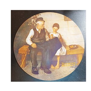 May include: A round ceramic plate featuring a painting of an older man and a young girl sitting on a wooden bench. The man is wearing a dark blue vest and pants, while the girl is wearing a white dress. The painting is signed "Norman Rockwell" in the bottom right corner.