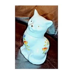 May include: A white ceramic cat figurine with a floral design. The cat is sitting with its head tilted to the side and its tail curled around its body.