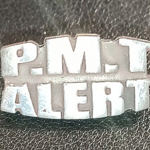Pode incluir: Um alfinete de metal prateado com o texto "P.M.T. ALERT" em letras em relevo.