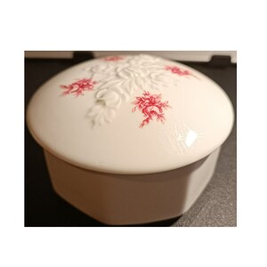 May include: White porcelain trinket box with a hexagonal base and a domed lid. The lid features a raised floral design with pink accents.