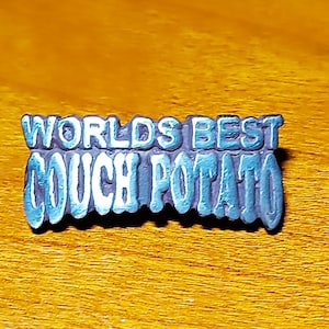 Könnte beinhalten: Eine blaue und weiße Metallnadel mit dem Text "Worlds Best Couch Potato" in einer stilisierten Schrift.