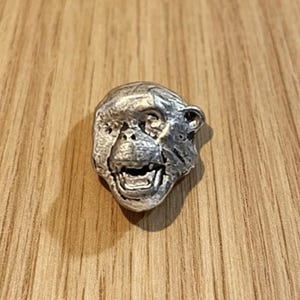 May include: A small, silver-toned metal pin shaped like the head of a primate. The piece features a textured surface with a detailed facial expression, including a visible mouth, nose, and eyes, capturing the likeness of a monkey or ape.