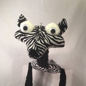 May include: A black and white zebra puppet with large googly eyes. The puppet has long black legs with black fur and zebra print feet.