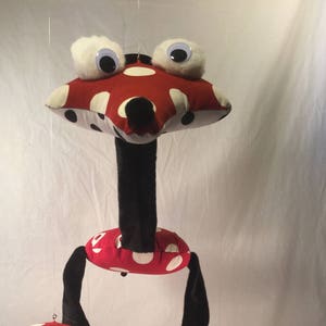 May include: A red and white polka dot mushroom-shaped plush toy with black legs and large white googly eyes. The toy is hanging from the ceiling.
