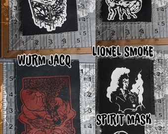 May include: Four black fabric patches with white designs. The patches are labeled "CUBE", "BOTFLY", "WURM JACQ", and "SPIRIT MASK". The "CUBE" patch features a cube with eyes and tentacles. The "BOTFLY" patch features a fly with wings. The "WURM JACQ" patch features a red and black illustration of a creature with a skull and the text "Nazi Devourer". The "SPIRIT MASK" patch features a skull with a mask and the text "LIONEL SMOKE".