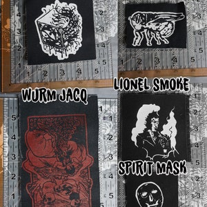 Könnte beinhalten: Vier schwarze Stoffpatches mit weißen Designs. Die Patches sind mit "CUBE", "BOTFLY", "WURM JACQ" und "SPIRIT MASK" beschriftet. Der "CUBE"-Patch zeigt einen Würfel mit Augen und Tentakeln. Der "BOTFLY"-Patch zeigt eine Fliege mit Flügeln. Der "WURM JACQ"-Patch zeigt eine rot-schwarze Illustration einer Kreatur mit einem Schädel und dem Text "Nazi Devourer". Der "SPIRIT MASK"-Patch zeigt einen Schädel mit einer Maske und dem Text "LIONEL SMOKE".