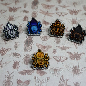 May include: Five colorful spider brooches with different designs. The brooches are made of resin and have a glossy finish. The spiders are in various colors, including blue, yellow, black, and white.