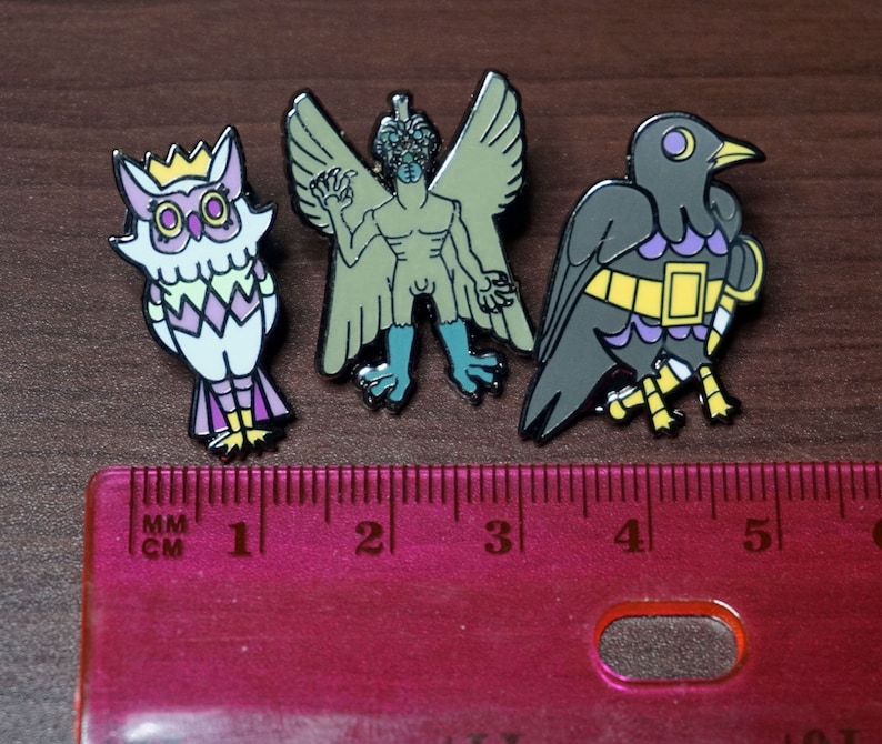 May include: Three enamel pins featuring a green and blue winged creature, a purple and white owl, and a black and yellow bird. The pins are arranged on a pink ruler with centimeter and inch markings.