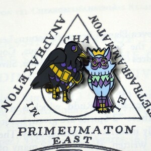 May include: Two enamel pins featuring a black crow and a blue owl. The crow has a gold band around its chest and purple accents. The owl has a gold crown and a purple, green, and white patterned design. The pins are set against a white background with a black triangle and text that reads "IMPRIMATUMON EAST" and "TETRAGRAMMATON".