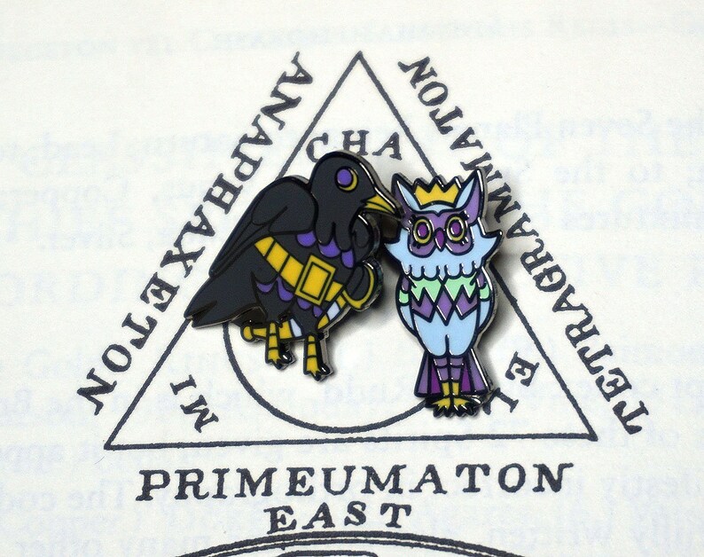 May include: Two enamel pins featuring a black crow and a blue owl. The crow has a gold band around its chest and purple accents. The owl has a gold crown and a purple, green, and white patterned design. The pins are set against a white background with a black triangle and text that reads "IMPRIMATUMON EAST" and "TETRAGRAMMATON".
