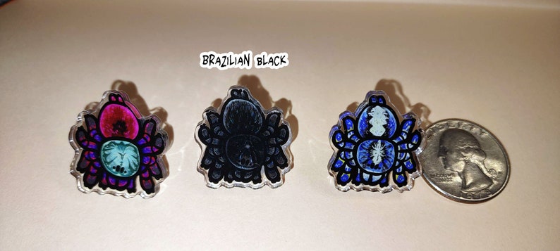 May include: Three clear acrylic pins featuring a spider design. The spiders are in different colors: pink, black, and blue. The pins are about the size of a US quarter.
