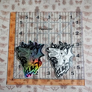May include: Two iridescent holographic stickers of a dragon with its mouth open, one in black and white and the other with rainbow colors. The stickers are on a ruler with inch markings.
