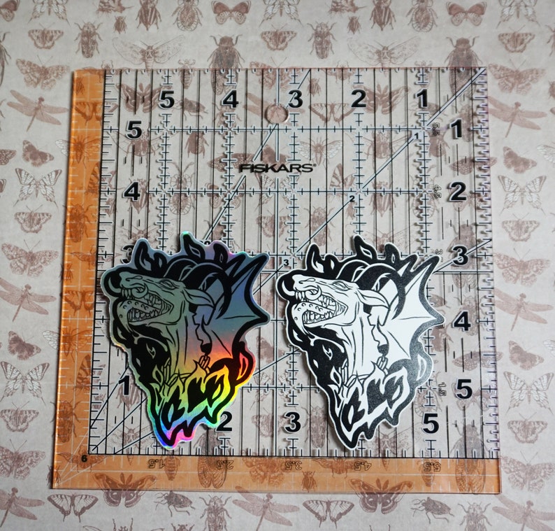 May include: Two iridescent holographic stickers of a dragon with its mouth open, one in black and white and the other with rainbow colors. The stickers are on a ruler with inch markings.