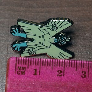 May include: A green and blue enamel pin of a winged creature with three eyes and clawed hands. The pin is about 2 inches long and is measured against a ruler.