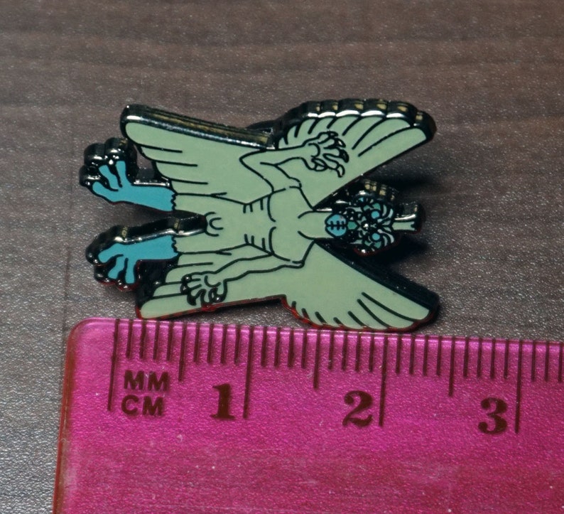 May include: A green and blue enamel pin of a winged creature with three eyes and clawed hands. The pin is about 2 inches long and is measured against a ruler.