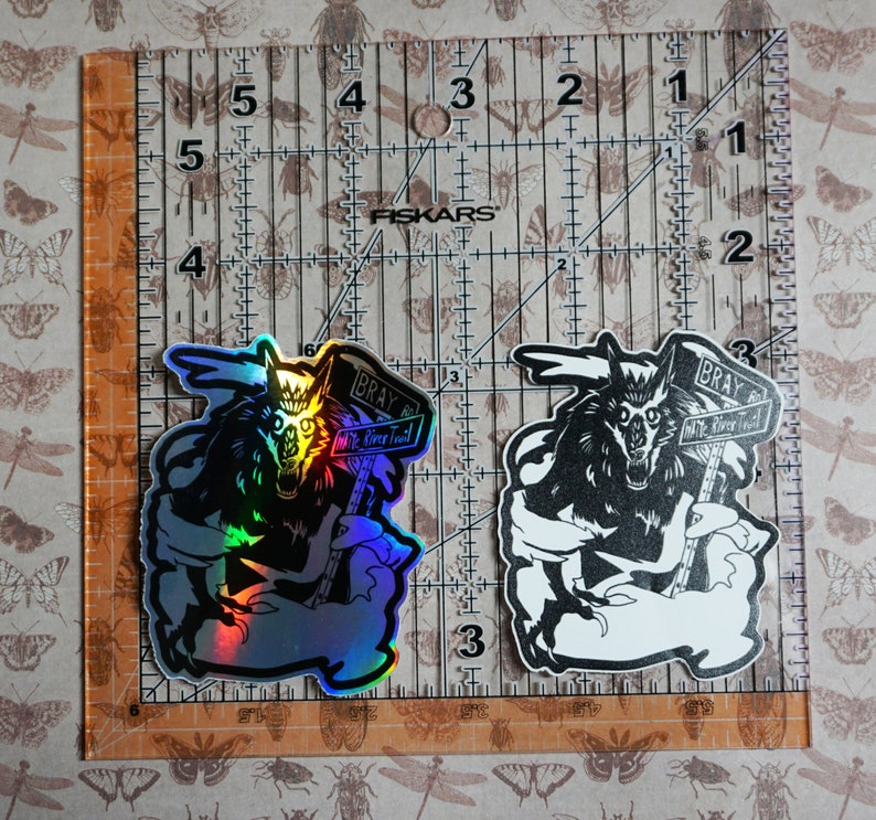 May include: Two stickers of a wolf holding a sign that says "Bray Wolf River Trail". One sticker is a holographic silver color and the other is black and white.