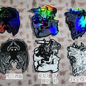 May include: Six holographic stickers featuring black and white illustrations of mythical creatures. The stickers are on a background of brown paper with a pattern of insects. The stickers are labeled "Mothman", "Beast of Bray Rd", and "Jersey Devil".